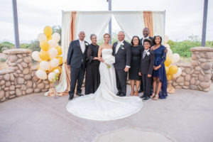The bride and groom, her parents, her sister, brother in-law, niece and nephew standing in from the ceremony backdrop with a cluster of balloons and a tall clear vase with a large ivory rose standing up in the vase.